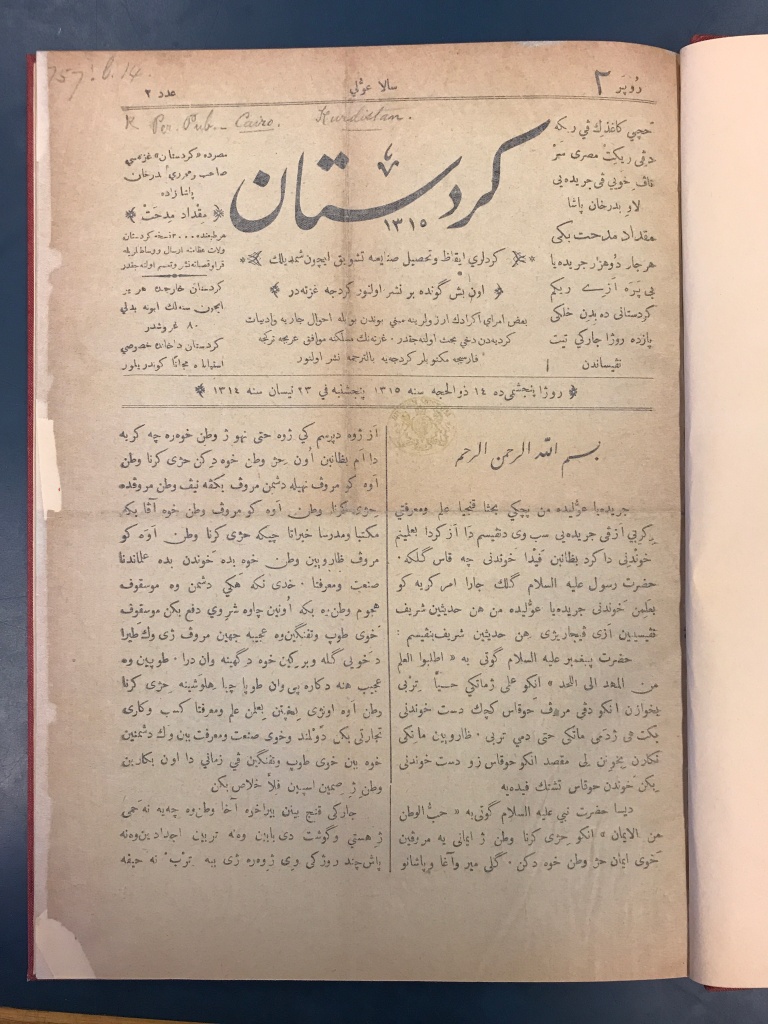 Cream-coloured page with text in Arabic script in black ink, with the masthead at the top in calligraphic script. A dark table top is visible in the margins. 